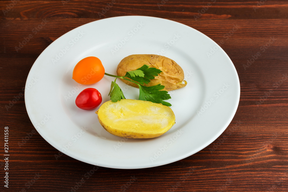 Slices of boiled potatoes, two tomatoes and a leaf of parsley, lie on a white round plate on a wooden brown table. Slices of boiled potatoes, two tomatoes and a leaf of parsley, lie on a white round plate on a wooden brown table.