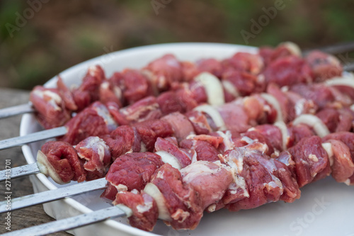 Five skewers with pieces of raw lamb with salt, black pepper and onion on a white round dish on an old wooden table, close up