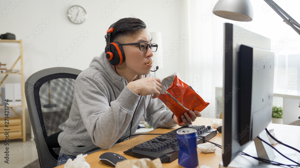 funny asian man with headphones plays video games on computer and eats snack from pack. japanese funny asian man with headphones plays video games on computer and eats snack from pack. japanese