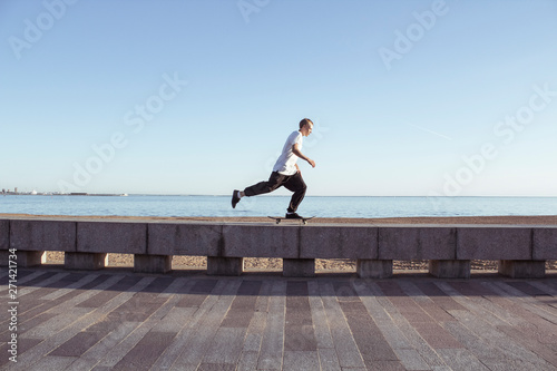 Skater rides on the parapet against the backdrop of the sea and the beach. Young european guy on a skateboard. Summer extreme sport skateboarding. Street style.