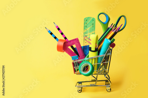 Back to school concept with shopping cart and colorful pencils, square ruler, scissors, clips, markers on pastel yellow backdrop. Flat lay, top view, copy space.
