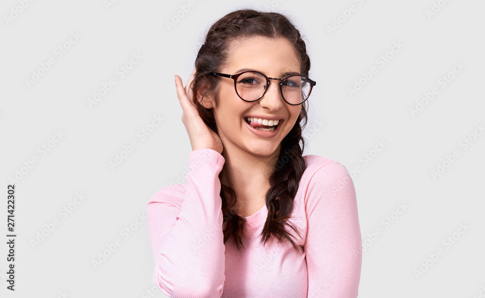 Portrait of funny young woman showing her tongue, blinking with an eye and wearing round trendy transparent spectacles while posing on white studio wall. Positive smiling female making grimace. People