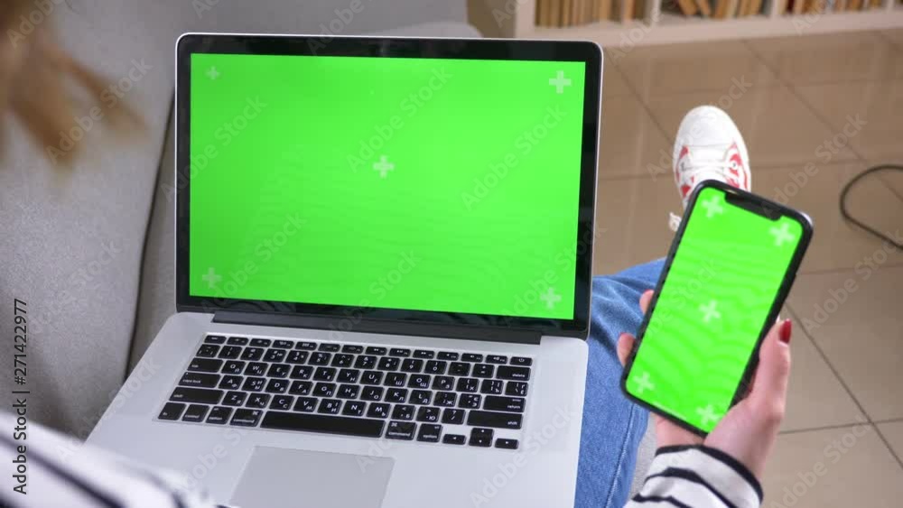 Closeup back view shoot of female using the laptop and phone with green screen sitting on the couch indoors in a cozy apartment
