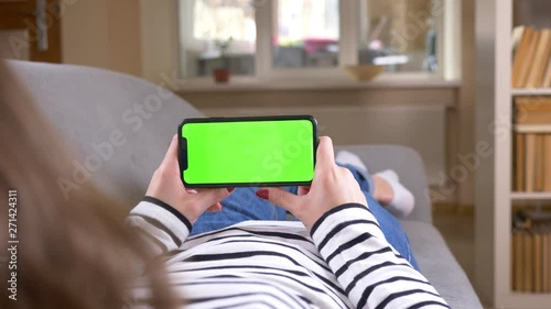 Closeup back view shoot of female using the phone with green screen holding it horizontally laying on the couch indoors in a cozy apartment