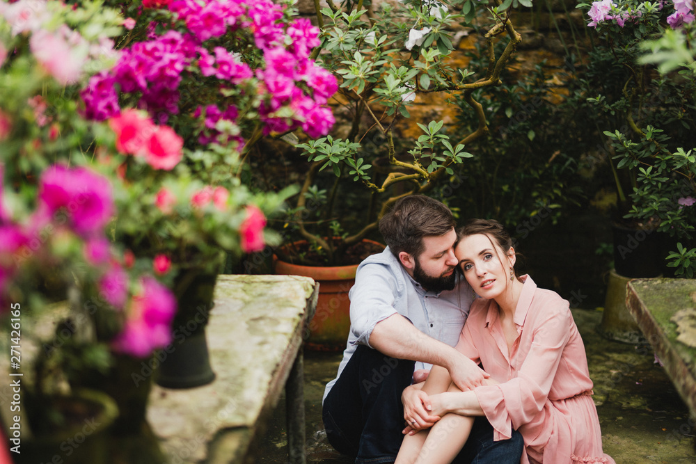 Fototapeta premium Young loving cheerful caucasian couple in glass greenhouse among colorful azalea flowers. Happy man and woman hugging and holding hands, spending time togehter.