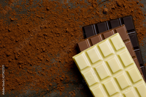 Obraz na plátně Slabs of white, milk and dark chocolate on top of each other on a black slate dusted with cocoa powder from above