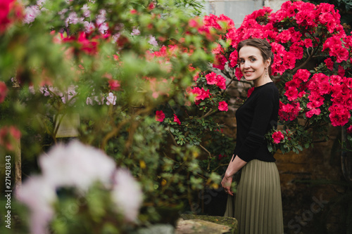 Young beautiful caucasian woman in glass greenhouse among colorful azalea flowers. Art portrait of a girl wearing classic fashionable clothes.