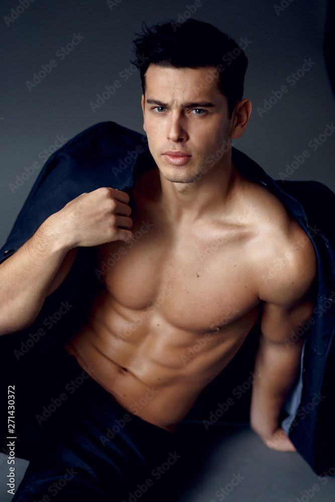 Muscular, young brunette man sitting and posing in fashionable suit on a naked torso, isolated on grey background. Muscular, young brunette man sitting and posing in fashionable suit on a naked torso, isolated on grey background.