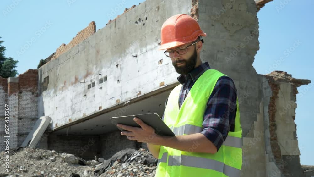 Bearded builder in a helmet and reflective vest uses a tablet on the background of a destroyed building, preparing for demolition and construction Bearded builder in a helmet and reflective vest uses a tablet on the background of a destroyed building, preparing for demolition and construction