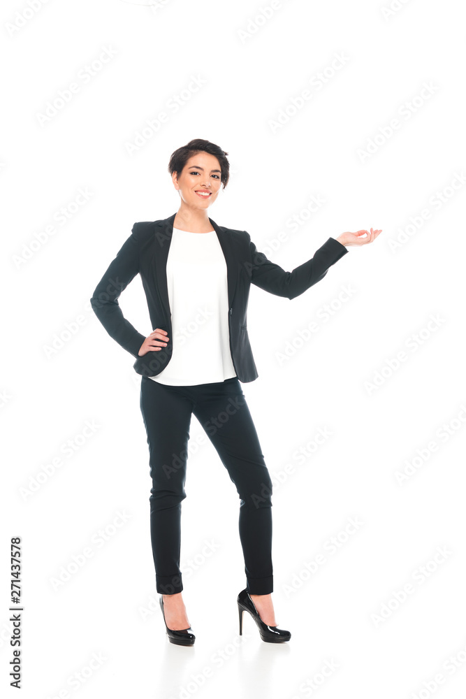 smiling mixed race businesswoman holding hand on hip and gesturing while looking at camera isolated on white smiling mixed race businesswoman holding hand on hip and gesturing while looking at camera isolated on white