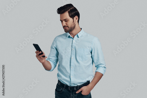 Always available. Handsome young man using smart phone and smiling while standing against grey background © G-Stock Faces