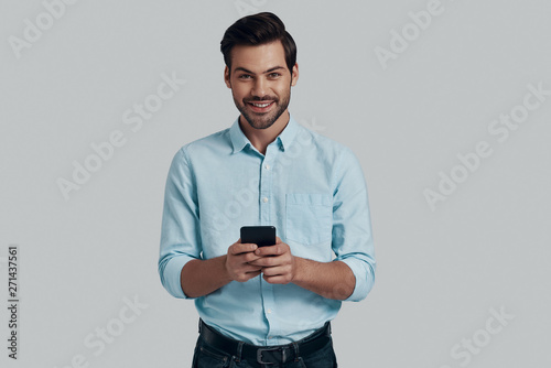 Typing message. Handsome young man using smart phone and looking at camera while standing against grey background © G-Stock Faces