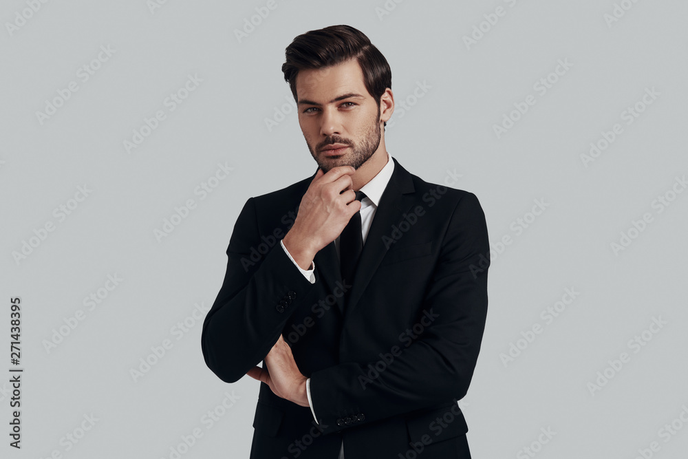 Pure elegance. Handsome young man in full suit keeping hand on chin and looking at camera while standing against grey background Pure elegance. Handsome young man in full suit keeping hand on chin and looking at camera while standing against grey background