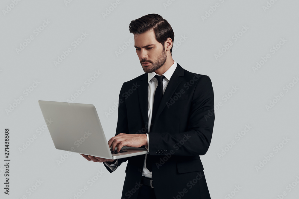 Working on the go. Handsome young man in full suit using laptop while standing against grey background Working on the go. Handsome young man in full suit using laptop while standing against grey background