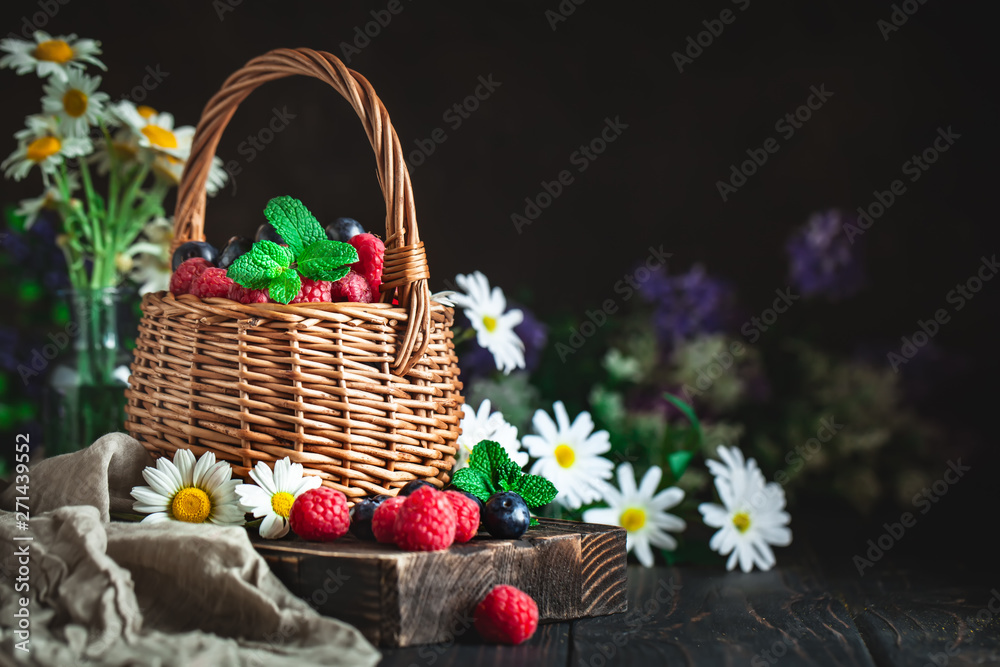 Raspberries and blueberries in a basket with chamomile and leaves on a dark background. Summer and healthy food concept. Background with copy space. Selective focus.