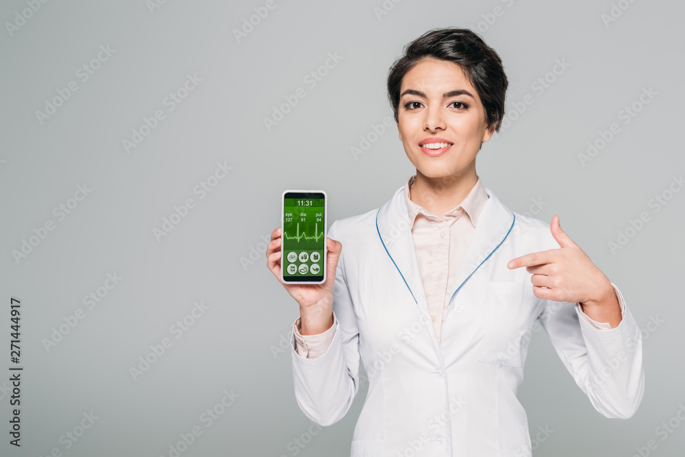 smiling mixed race doctor pointing with finger at heartbeat rate app on screen isolated on grey smiling mixed race doctor pointing with finger at heartbeat rate app on screen isolated on grey
