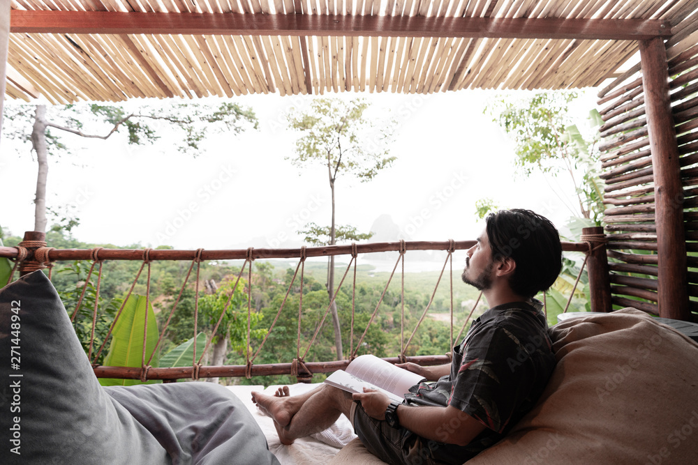 Asian men traveler wear black shirt while reading book looking forward to next trip and sitting on bean bag on resort balcony in relax mood vacation time/travel concept / resort mood / rejuvenate time