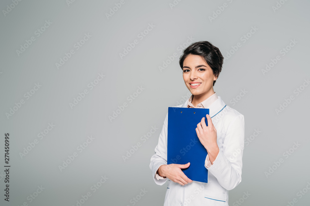 pretty mixed race doctor holding clipboard while smiling at camera isolated on grey pretty mixed race doctor holding clipboard while smiling at camera isolated on grey
