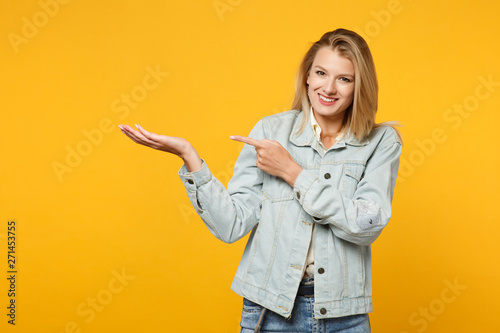 Portrait of smiling young woman in denim casual clothes looking camera, pointing index finger, hand aside isolated on yellow orange background in studio. People lifestyle concept. Mock up copy space. © ViDi Studio