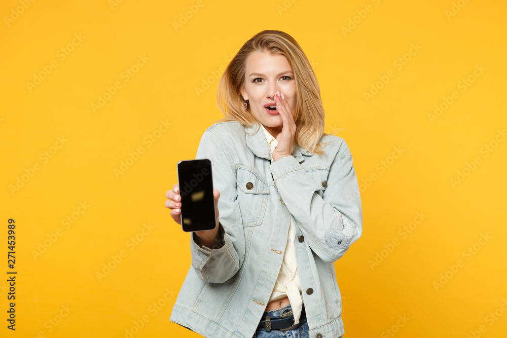 Excited young woman in denim clothes tell something with hand gesture, holding mobile phone with blank empty screen isolated on yellow orange background. People lifestyle concept. Mock up copy space.