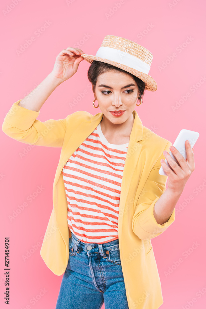 pretty mixed race woman touching straw hat while using smartphone isolated on pink pretty mixed race woman touching straw hat while using smartphone isolated on pink