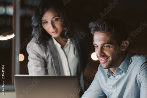 Smiling office colleagues using a laptop while working overtime together photo