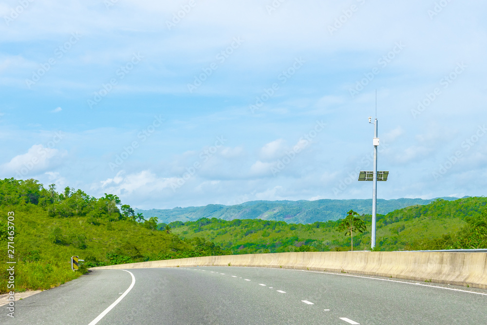 Energy efficient solar powered security system street camera on dual carriageway highway through mountain landscape. Vehicles drive on left hand side of this two lane asphalt road in St Ann, Jamaica.