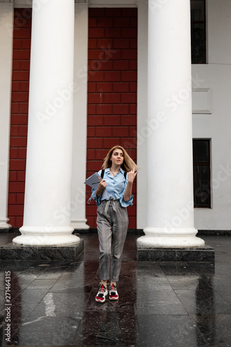 dreamy girl in a blue blouse with notebooks and a backpack stands against the background of the college. © mtrlin