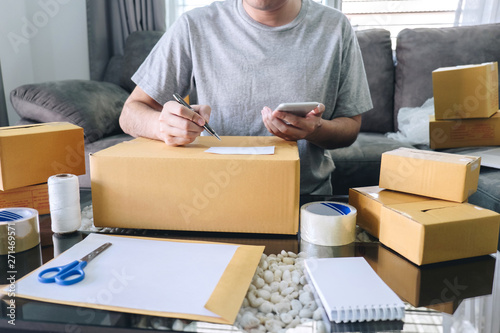 Young entrepreneur SME freelance man using smartphone receive order client and take note working with packaging sort box delivery online market on purchase order and preparing package product © Freedomz