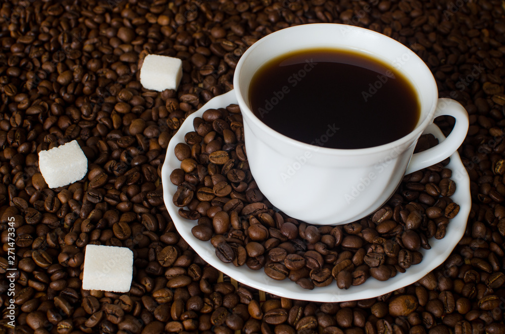 close-up of white Cup and saucer with black hot strong Americano coffee on a dark background of aromatic coffee beans with three pieces of white refined sugar. Flatlay close-up of white Cup and saucer with black hot strong Americano coffee on a dark background of aromatic coffee beans with three pieces of white refined sugar. Flatlay