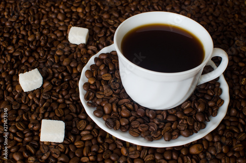 close-up of white Cup and saucer with black hot strong Americano coffee on a dark background of aromatic coffee beans with three pieces of white refined sugar. Flatlay