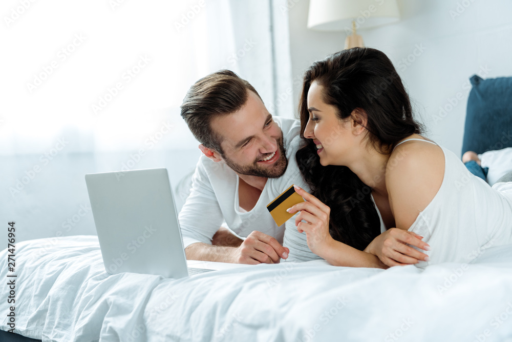 happy couple lying in bed near laptop while woman holding credit card, illustrative editorial happy couple lying in bed near laptop while woman holding credit card, illustrative editorial