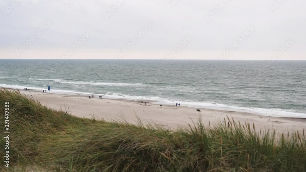 People in distance on sandy, overcast, Denmark beach. Grass on the dunes blowing in the wind as kite spins & flips along the coastline. People in distance on sandy, overcast, Denmark beach. Grass on the dunes blowing in the wind as kite spins & flips along the coastline.