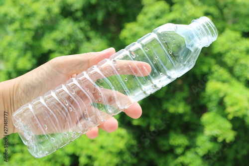 Man's hand holding an empty plastic bottle of drinking water against blurry green foliage