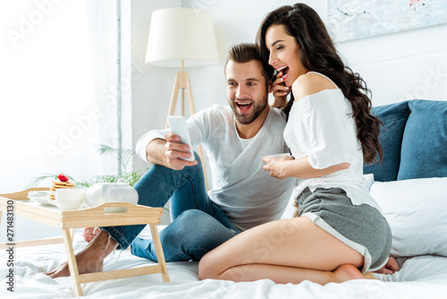 excited couple taking selfie near tray with pancakes and tea in bed