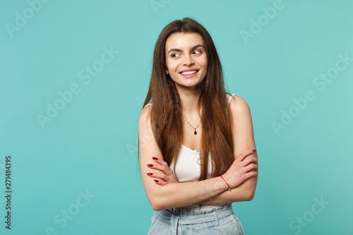 Portrait of smiling young woman in light casual clothes looking aside, holding hands crossed isolated on blue turquoise wall background. People sincere emotions, lifestyle concept. Mock up copy space.
