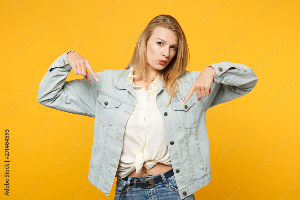 Fototapeta premium Portrait of pretty young woman in denim casual clothes looking camera, pointing index fingers down isolated on yellow orange wall background in studio. People lifestyle concept. Mock up copy space.
