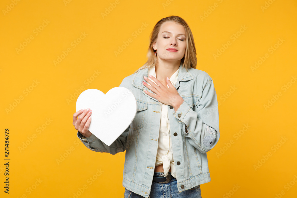 Pretty young woman in denim casual clothes keeping eyes closed, put hand on chest, hold white heart with copy space isolated on yellow orange background. People lifestyle concept. Mock up copy space. Pretty young woman in denim casual clothes keeping eyes closed, put hand on chest, hold white heart with copy space isolated on yellow orange background. People lifestyle concept. Mock up copy space.