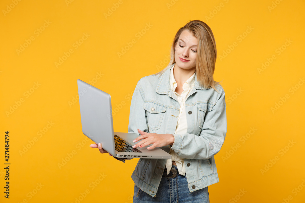 Portrait of smiling pretty young woman in denim casual clothes holding, using laptop pc computer isolated on yellow orange wall background in studio. People lifestyle concept. Mock up copy space.