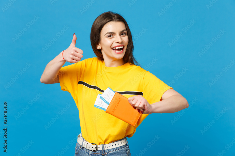 Cheerful young woman in vivid casual clothes looking camera, showing thumb up, holding passport, boarding pass ticket isolated on blue wall background. People lifestyle concept. Mock up copy space.