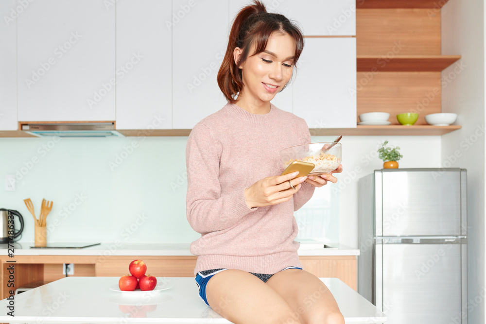 Asian young woman sitting on the table reading a message on her mobile phone and smiling while eating cornflakes with milk for breakfast