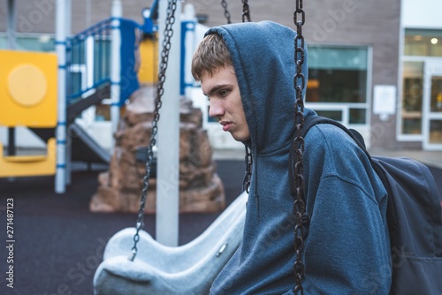 Sad teenager sitting on a swing outside of a school. He is reminiscing about when he was younger. photo