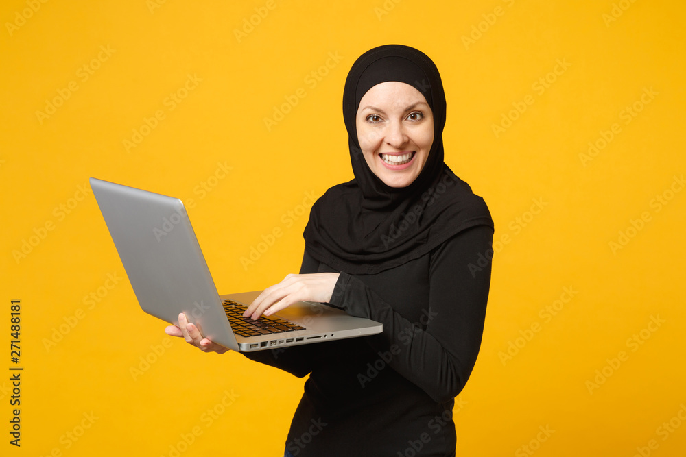Young arabian muslim employee woman in hijab black clothes hold and work laptop pc computer isolated on yellow wall background, studio portrait. People religious lifestyle concept. Mock up copy space. Young arabian muslim employee woman in hijab black clothes hold and work laptop pc computer isolated on yellow wall background, studio portrait. People religious lifestyle concept. Mock up copy space.