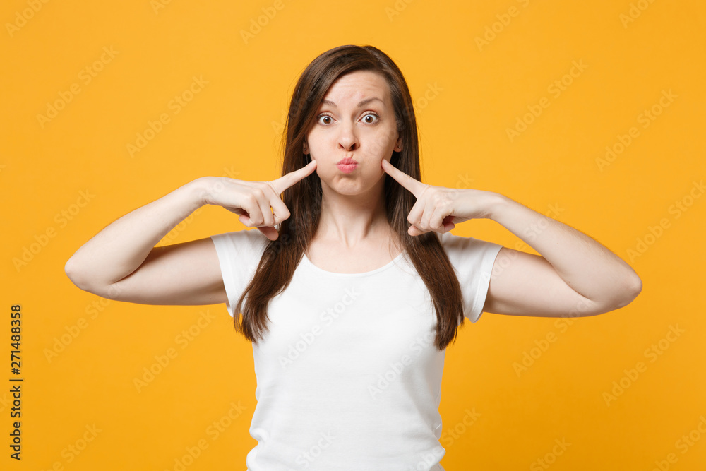 Fototapeta premium Portrait of funny young woman in white casual clothes pointing index fingers on blowing cheeks isolated on bright yellow orange wall background in studio. People lifestyle concept. Mock up copy space.