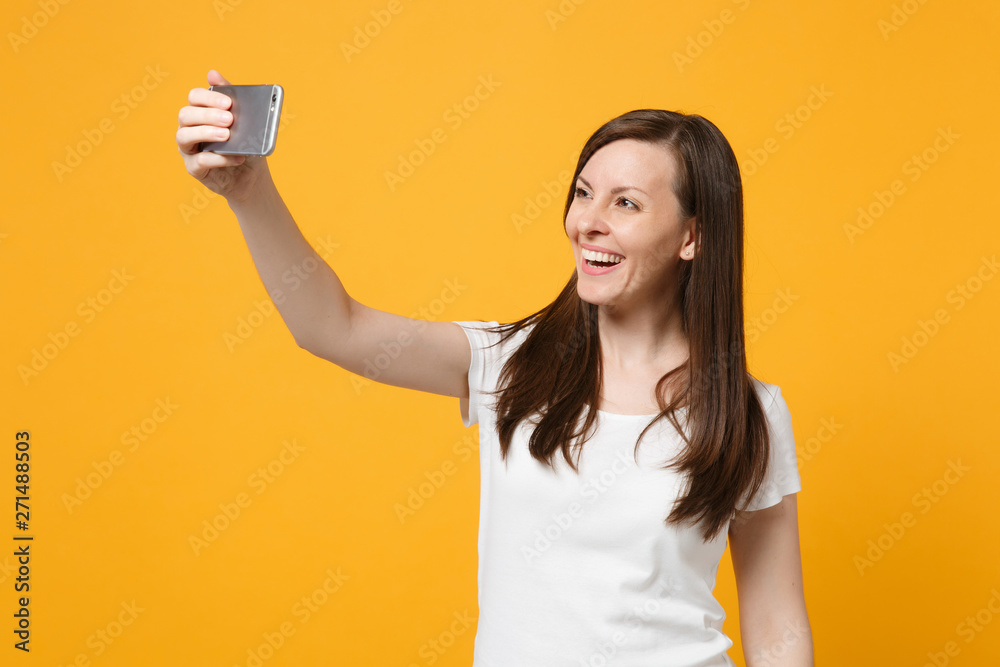 Portrait of cheerful young woman in white casual clothes doing selfie shot on mobile phone isolated on bright yellow orange wall background in studio. People lifestyle concept. Mock up copy space.