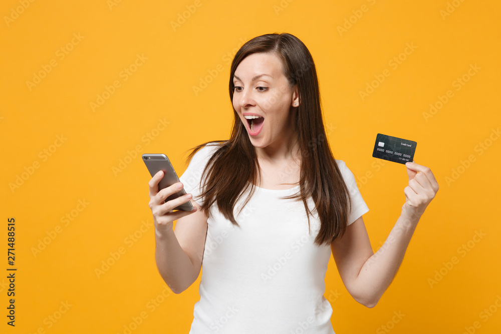 Excited young woman in white casual clothes keeping mouth open, using mobile phone, holding credit bank card isolated on bright yellow orange background. People lifestyle concept. Mock up copy space. - obrazy, fototapety, plakaty 