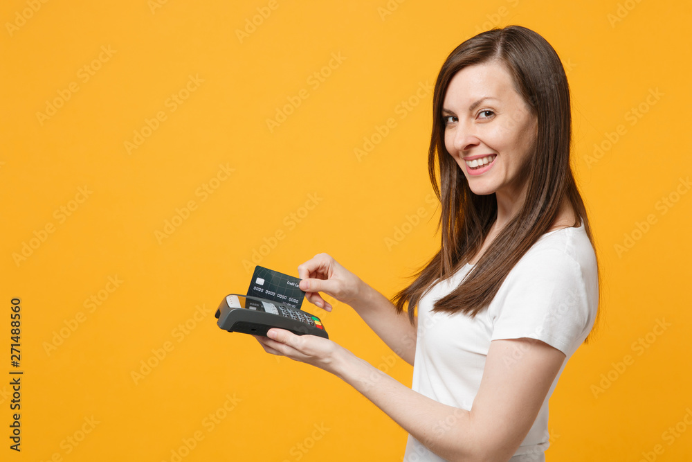 Smiling young woman holding wireless modern bank payment terminal to process and acquire credit card payments isolated on yellow orange wall background. People lifestyle concept. Mock up copy space. Smiling young woman holding wireless modern bank payment terminal to process and acquire credit card payments isolated on yellow orange wall background. People lifestyle concept. Mock up copy space.