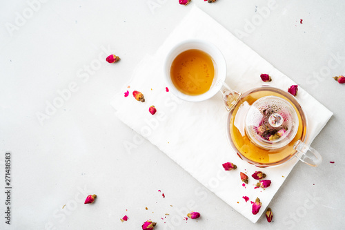 Top view of two white cups and transparent teapot with herbal tea of dried pink roses buds over textile napkin on gray background with copy space. Brewing and Drinking tea.