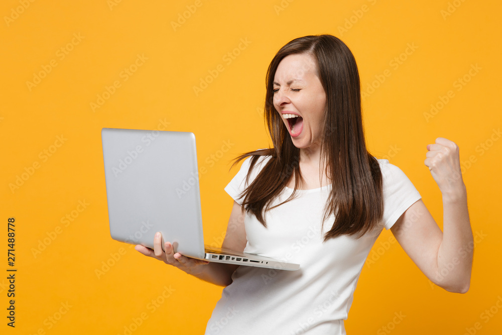 Screaming young woman in white casual clothes keeping eyes closed, doing winner gesture hold laptop pc computer isolated on yellow orange wall background. People lifestyle concept. Mock up copy space. Screaming young woman in white casual clothes keeping eyes closed, doing winner gesture hold laptop pc computer isolated on yellow orange wall background. People lifestyle concept. Mock up copy space.