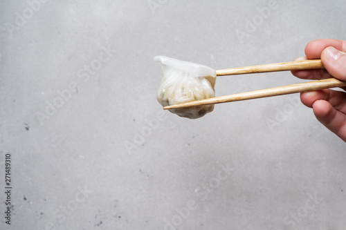 Close-up with a hand that holds a traditional Chinese dumpling steamed by Dim Sum with Har Gow on chopsticks. Asian food background.
